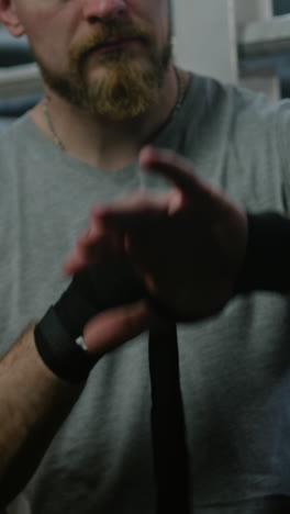 Adult-man-sits-near-boxing-ring-and-wraps-his-hands-with-bandage-for-martial-arts.-Athlete-prepares-to-fight,-competition-or-training-in-box-gym.-Boxer-performs-shadow-boxing-at-background.-Vertical-shot